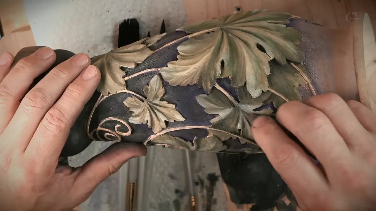 Finished vase held in both hands, olive green leaves standing off a near black ground with copper stems