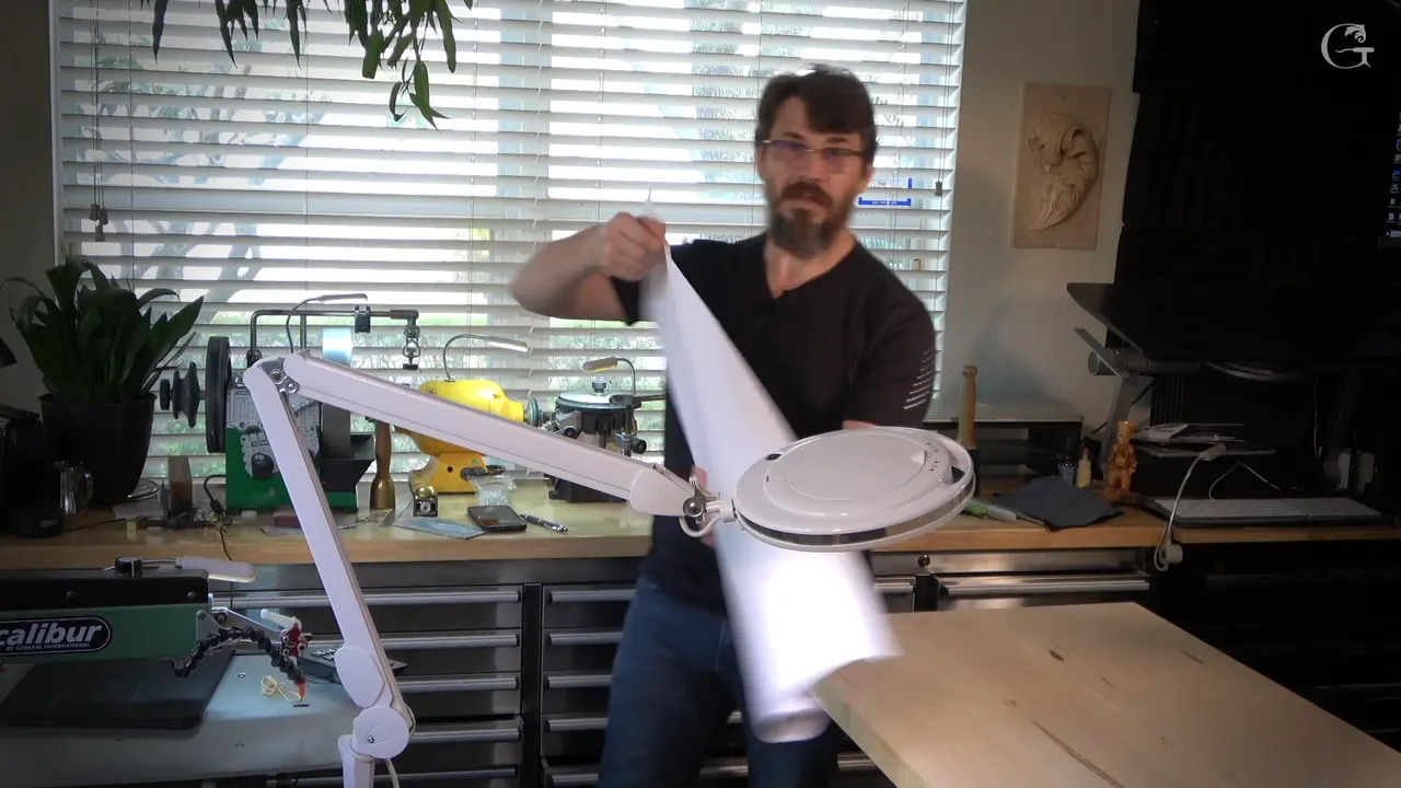 Alexander unrolls a long printed paper pattern across his bench, magnifier lamp and scroll saw behind him