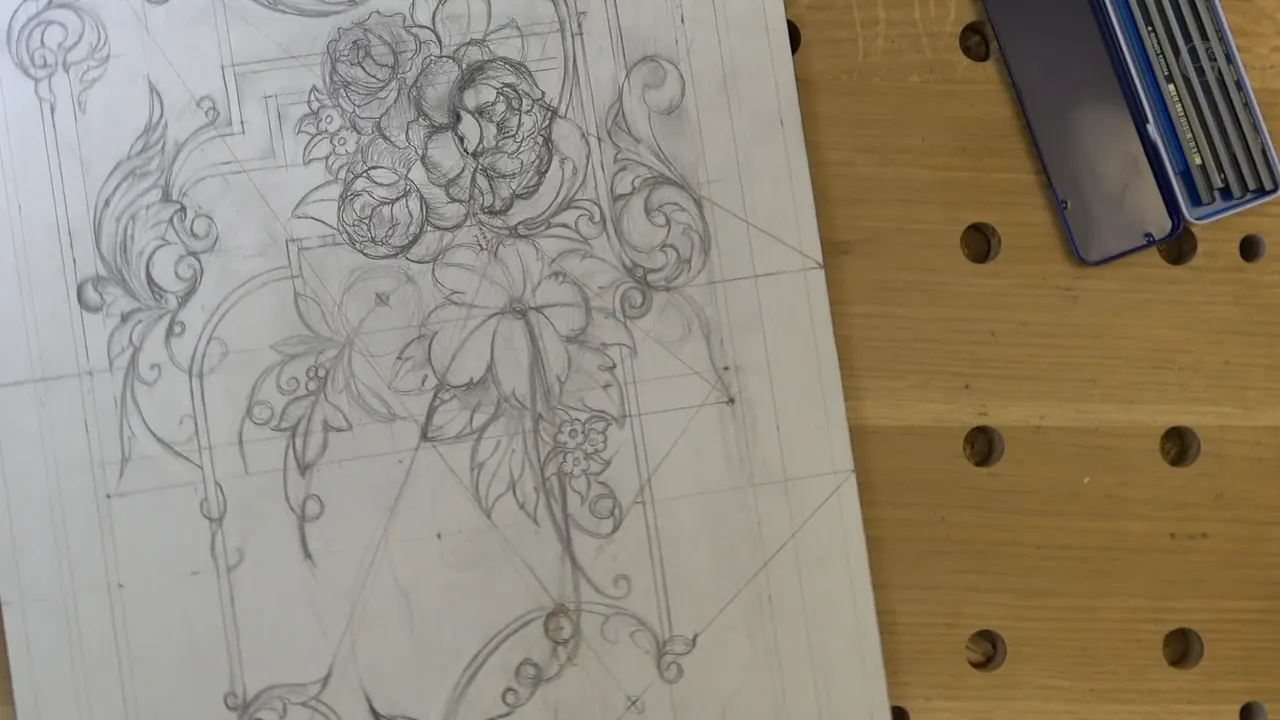 Full-size panel drawing pinned flat on the bench, a tin of pencils open beside it