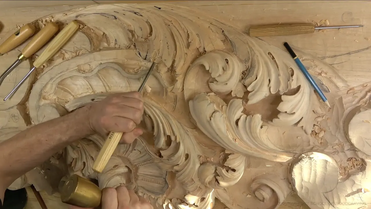 Seen from above, the carved board, one hand at work near a shell, a brass mallet and gouges lying nearby.