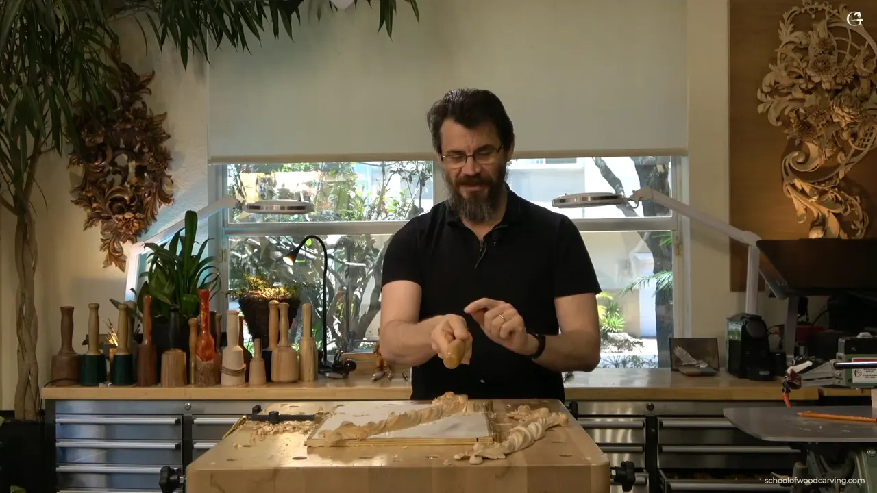 At the bench with the carved rosette held up in both hands and the tool pointing back toward him.