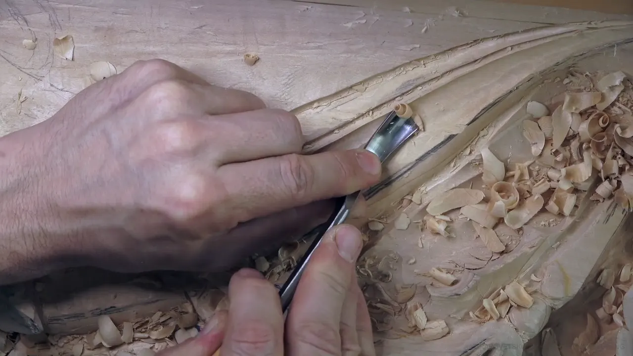 A curved gouge in both hands, carving the concave of the molding.
