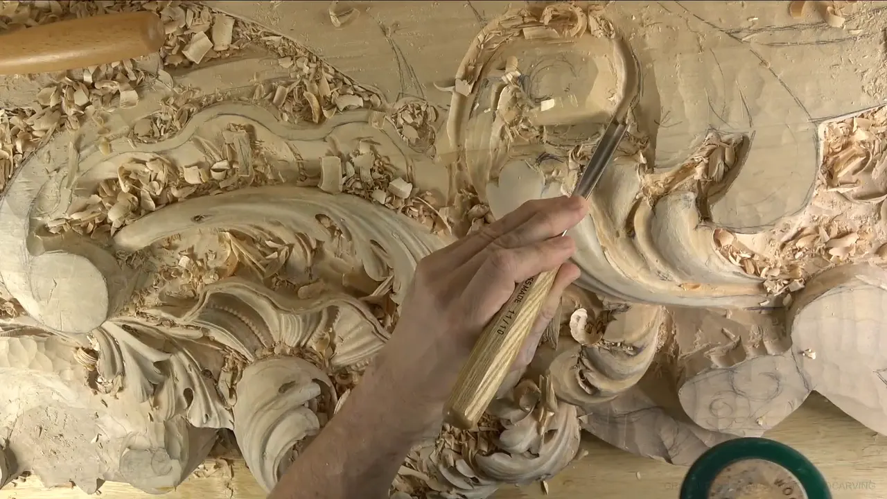 Wide view of the board, carved acanthus leaves at left, one hand working a gouge in at right.