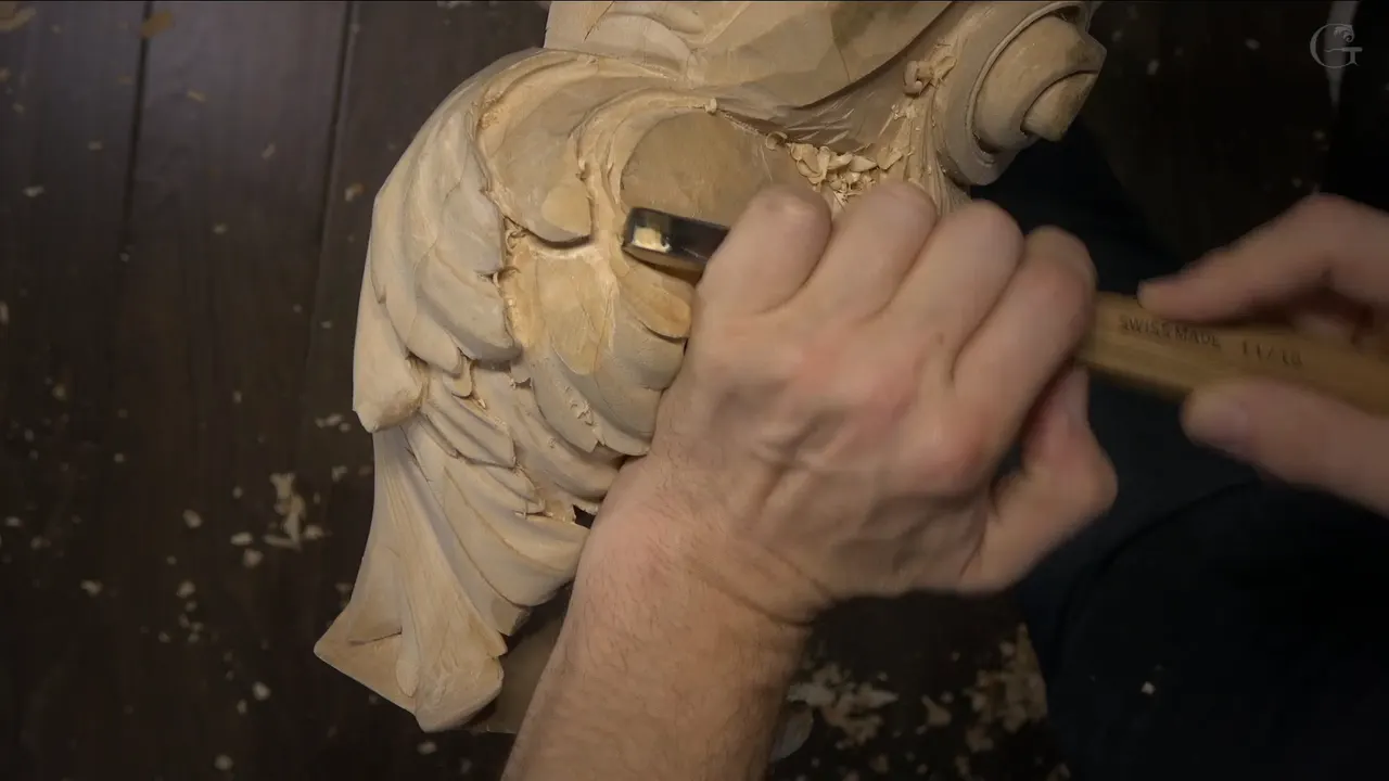 Two hands push a straight gouge into the smooth middle of the carving, leaves close on the left.