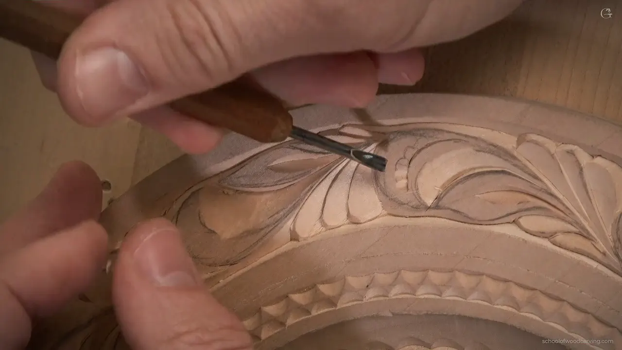 Fingers steer a tiny gouge with a dark handle into a carved leaf on the rim of the plate