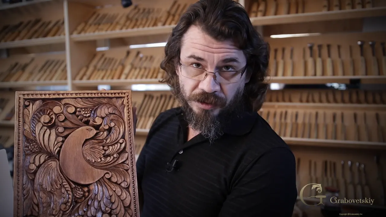 Alexander holding up the finished board, carved, stained with walnut shells and oiled, in front of his tool rack.