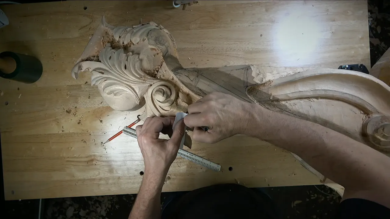 Two hands hold a small piece of paper against the carved edge, a triangular ruler and a red pencil below