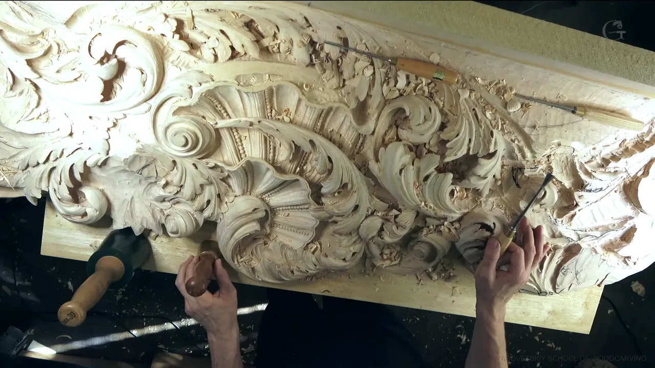 Whole carved ceiling panel on the bench, gouges lying on top, both hands working near the right end.