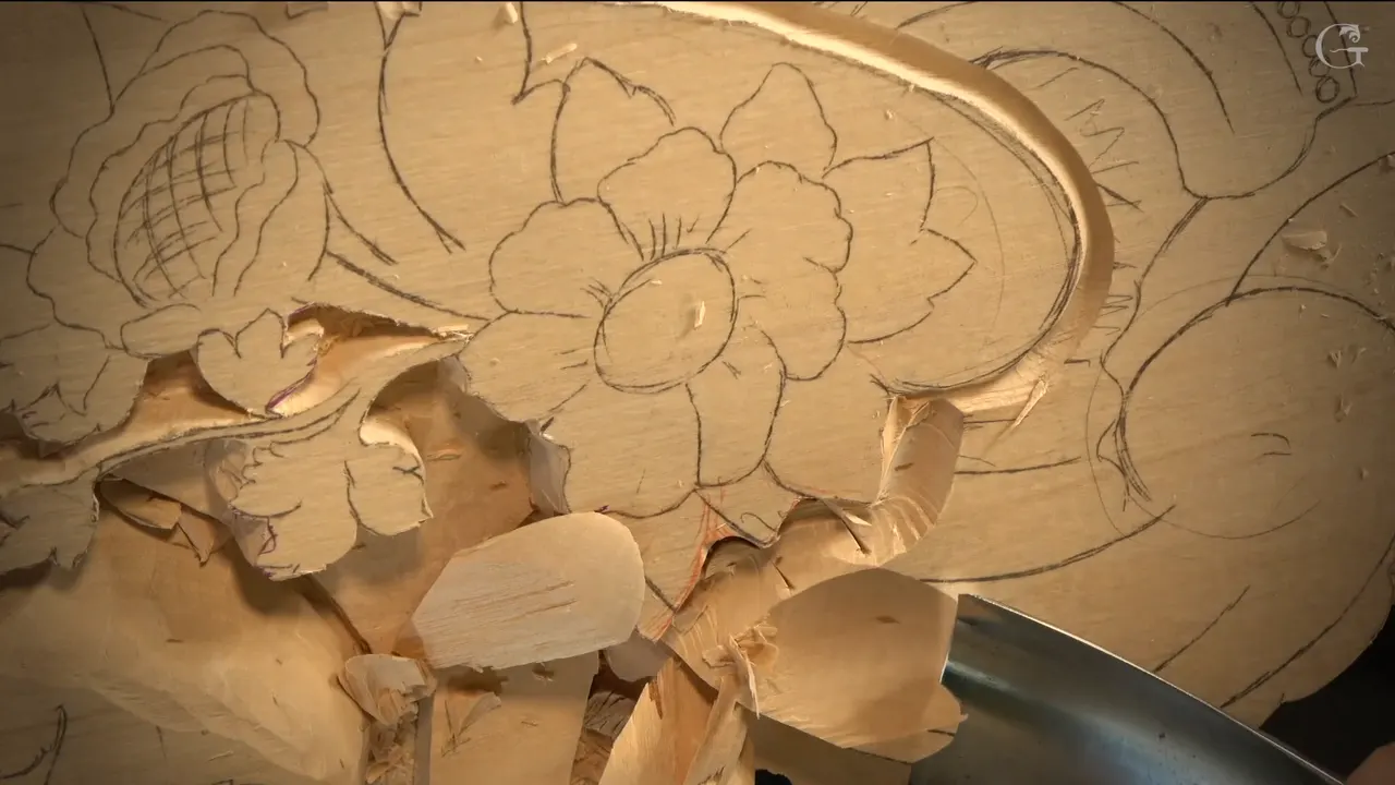 A very wide gouge blade pushes into the lowered wood, big chips lying loose beside a drawn flower.