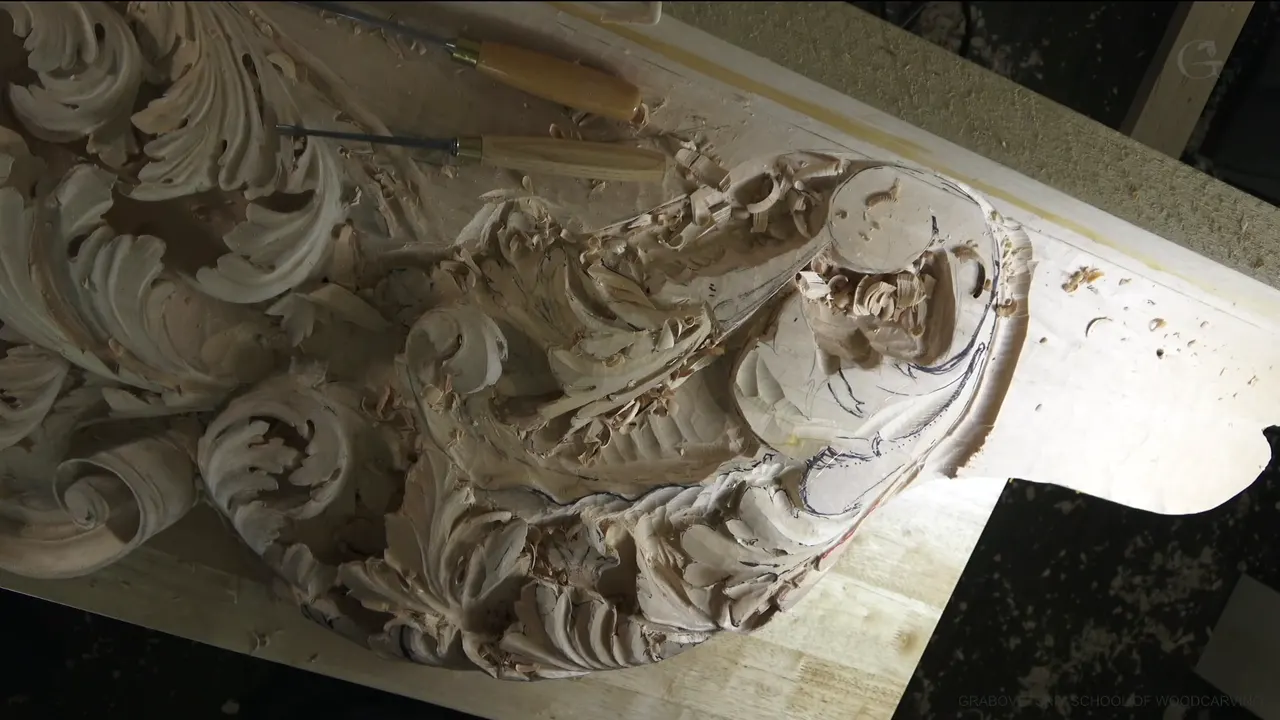 A section of the carved ceiling panel seen from above, deep shadows under the leaves and two gouges lying on the board.