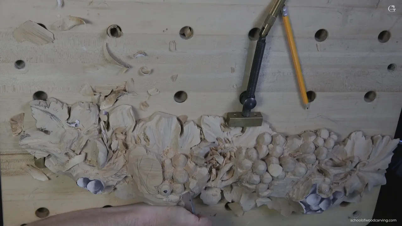 Whole panel clamped to the bench, both grape clusters roughed in, a pencil and a gouge lying beside the work