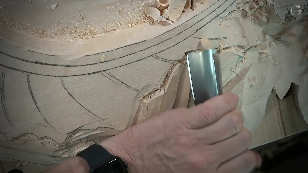 Huge U gouge held upright against the edge of the carved shape, with fingers and a watch below it