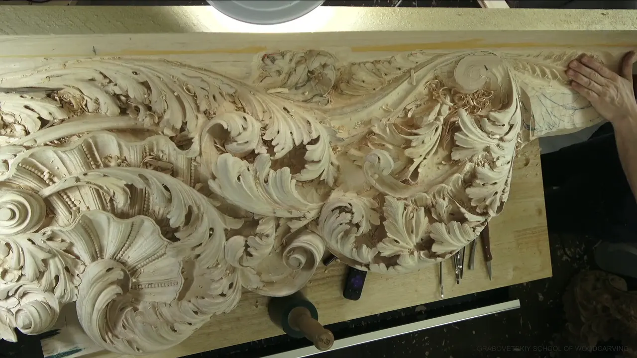 A long ceiling board of carved acanthus scrolls and shells, with tools lying on the bench below.
