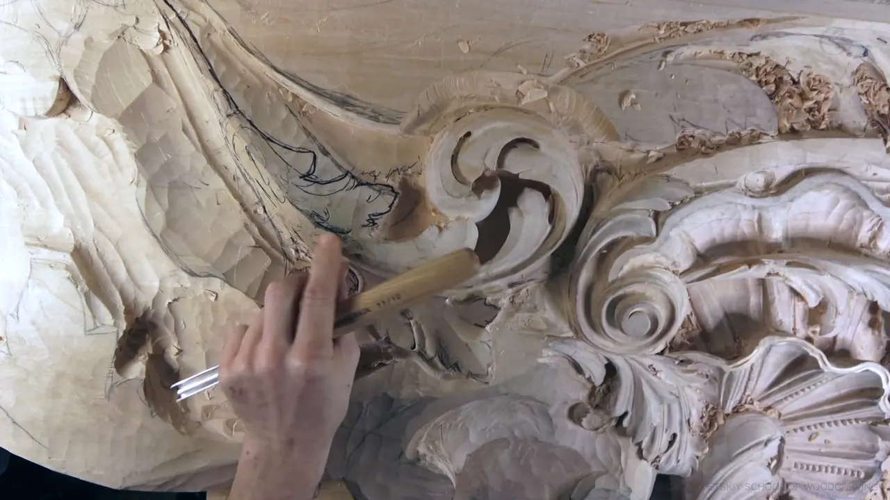 Overhead view of the carved scrolls and shell, a hand guiding a gouge along a dark line on the rough leaf.