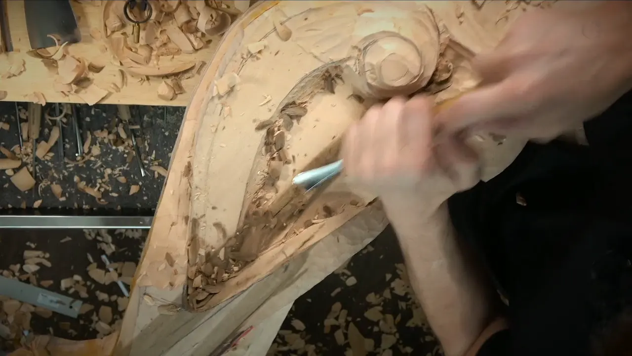 Both hands drive a gouge into the long hollow beside the carved scroll