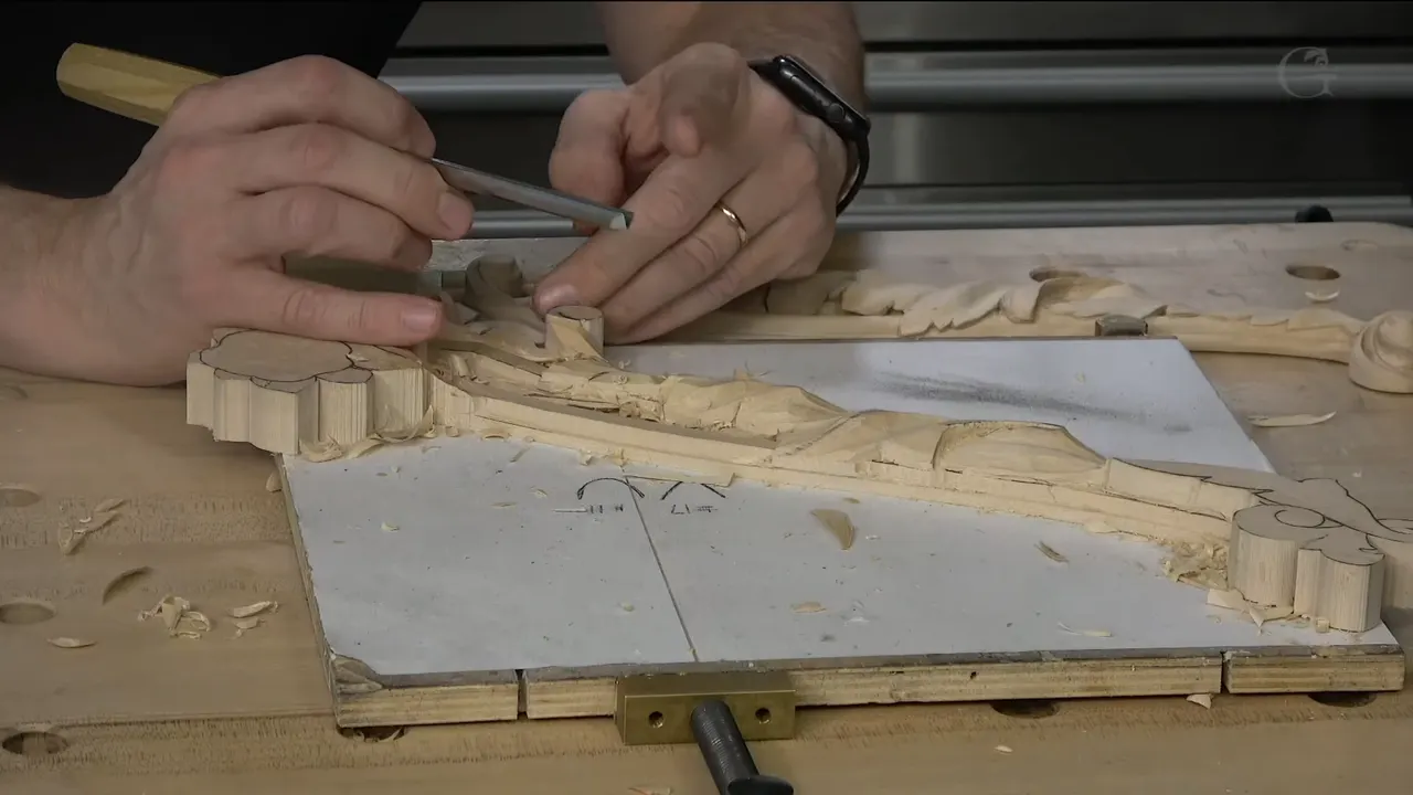 Both hands work the long carving on its white board, clamped at the front edge of the bench.