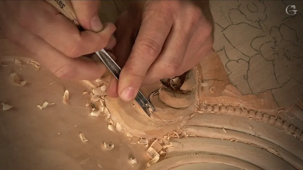 Two hands guide a slim gouge into a carved scroll, curls of wood lying around it