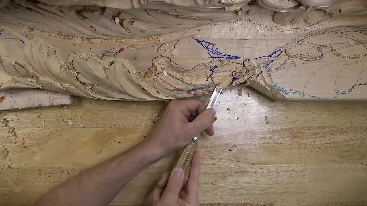 Two hands hold a gouge low against a thin carved section, with blue and red marker lines drawn above it