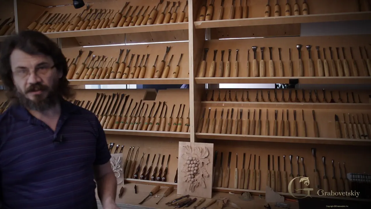 Alexander in front of five shelves of carving tools, a carved grape panel propped against the bench beside him.