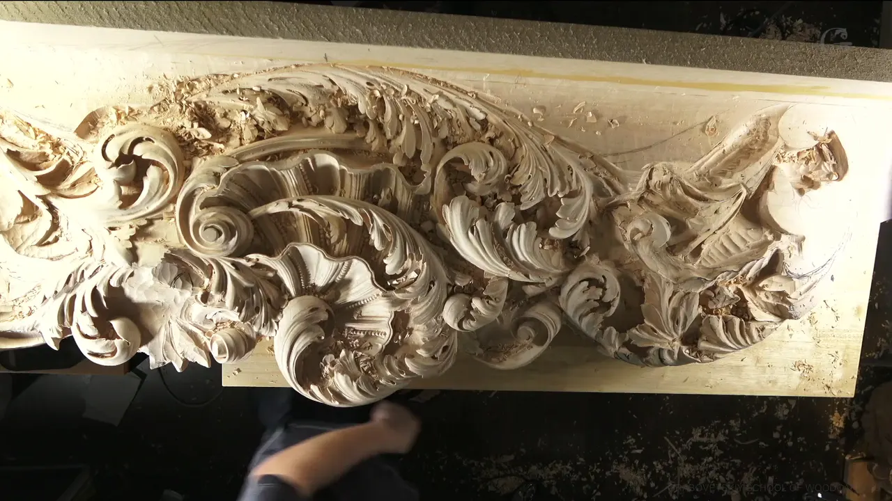 A long ceiling board seen from above, acanthus scrolls carved deep along it and one arm resting below.
