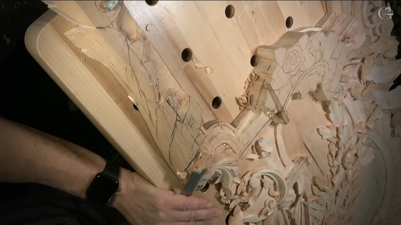 Seen from the side, a hand works a small tool into the carving below the flowers