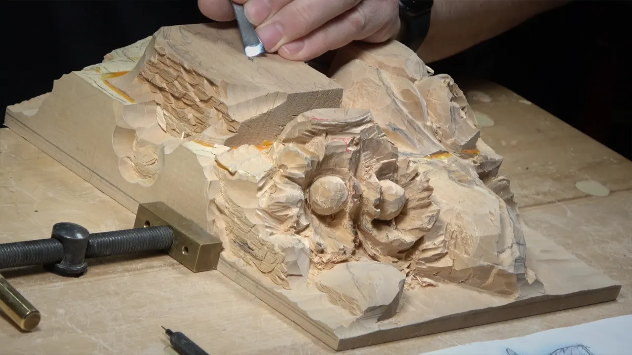 Hand with a narrow gouge on the tall block, two round nuts lower on the panel