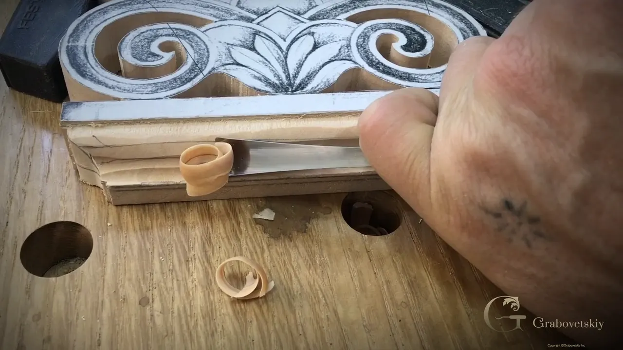 A gouge rides the stepped base of the acroterion and lifts one pale curl of basswood off the edge