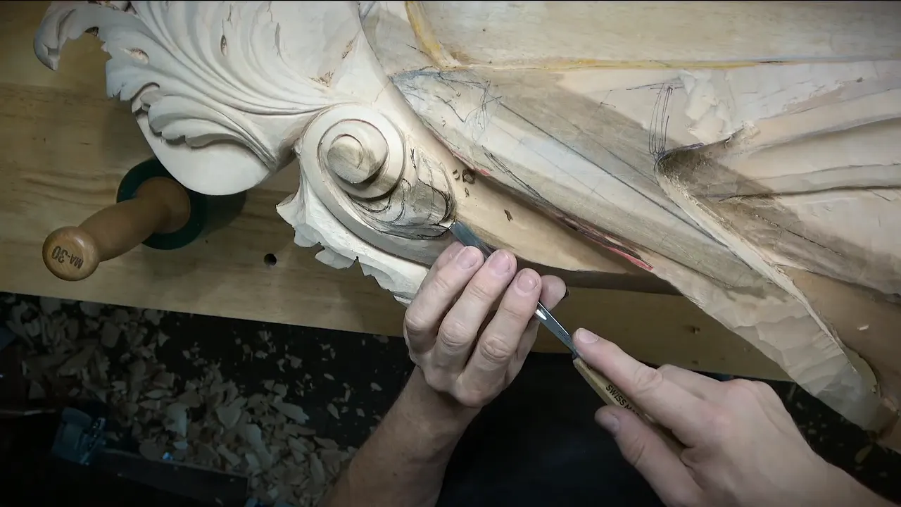 Two hands work a gouge into the curl of a large carved volute.