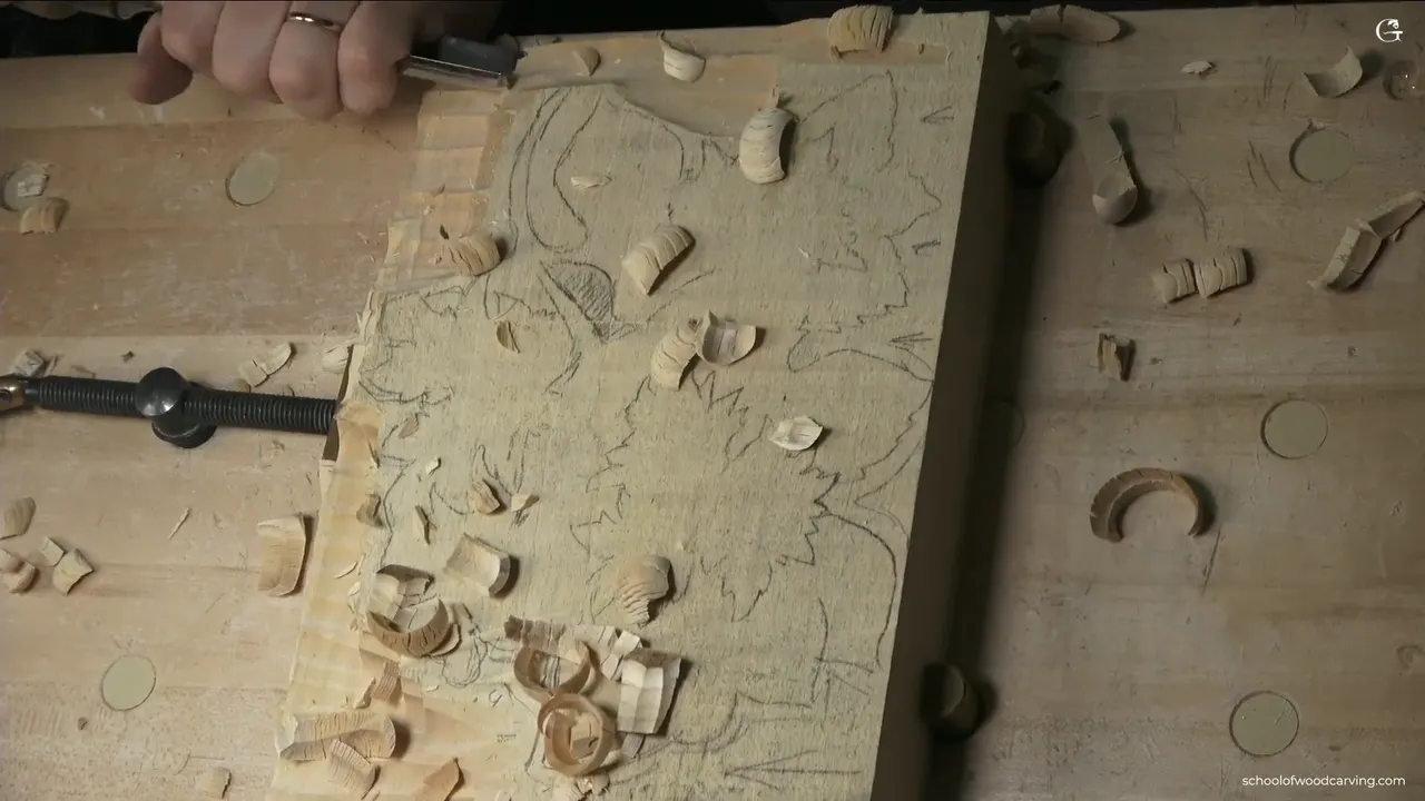 A hand on the gouge at the corner of the traced basswood panel, chips scattered across the top