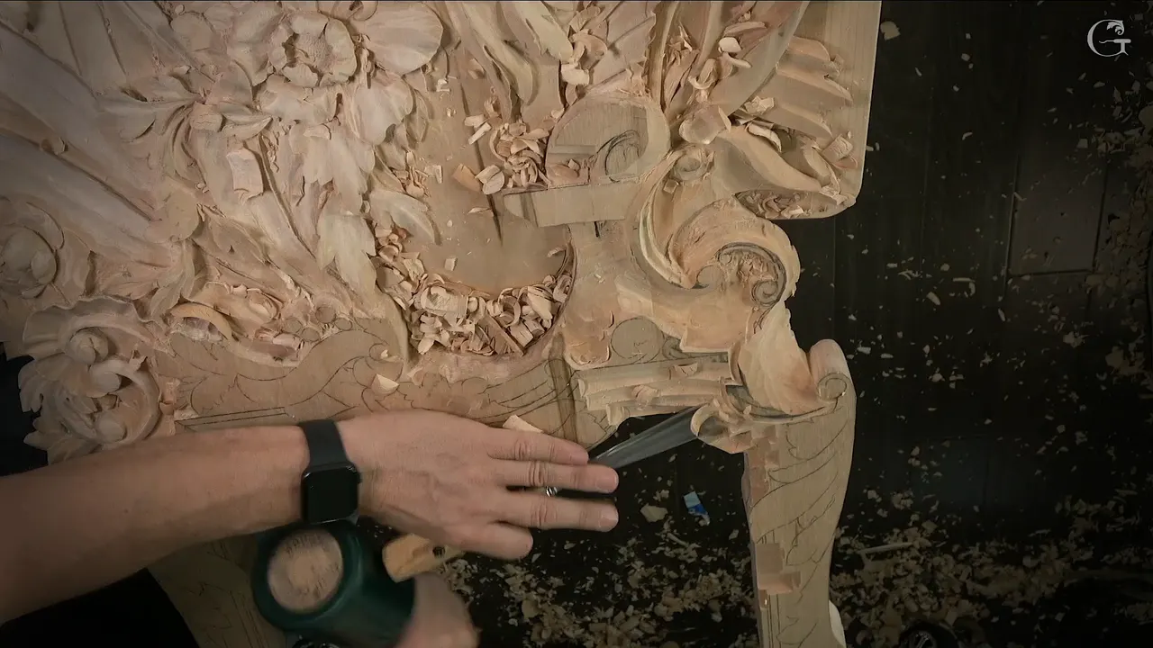 Green mallet in one hand, a gouge set against the rim of the carved panel in the other