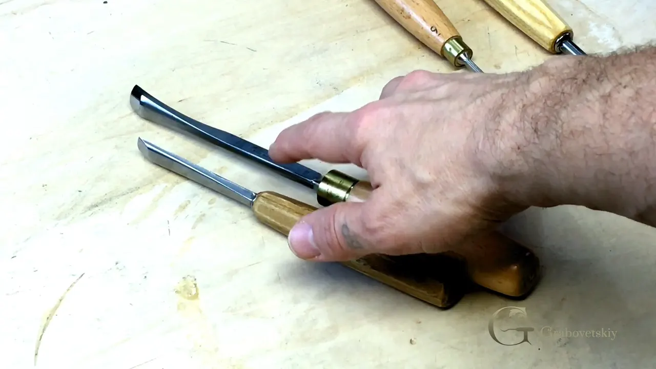 Two gouges laid side by side on a pale board, a hand resting on the handle of one