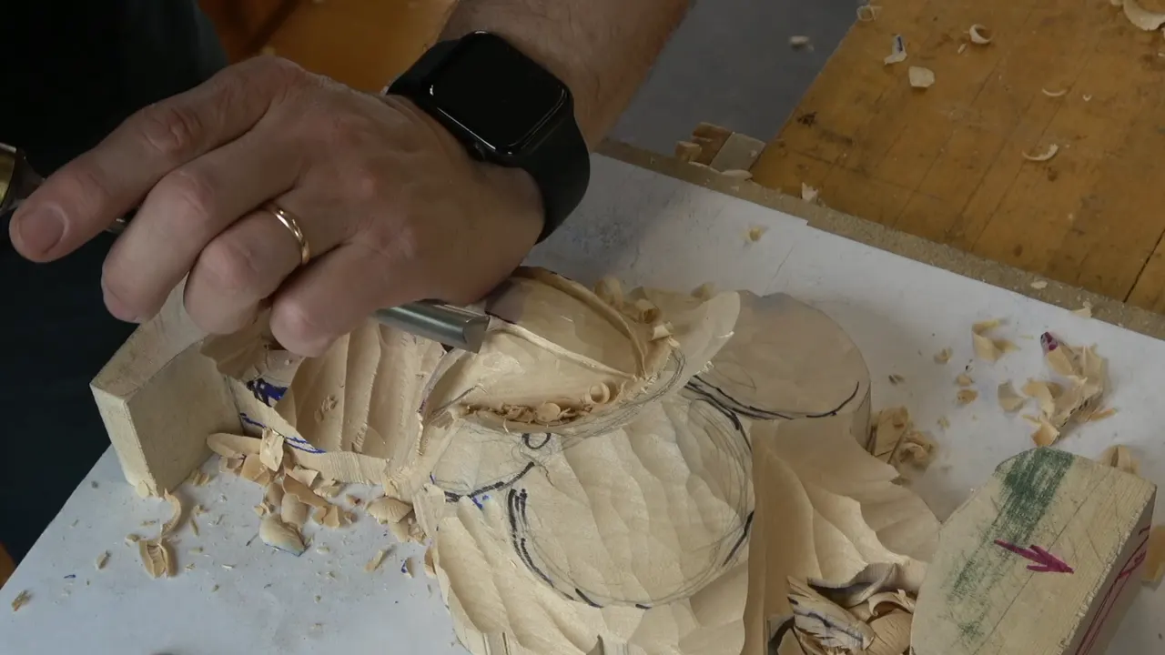 A hand with a watch drives a gouge into a pale block, chips all over a white sheet.