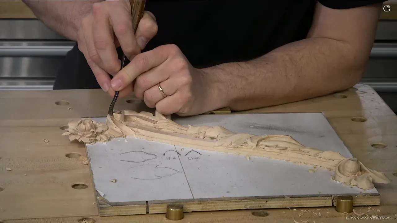 Both hands guide a gouge along a carved molding, a flower at its far end