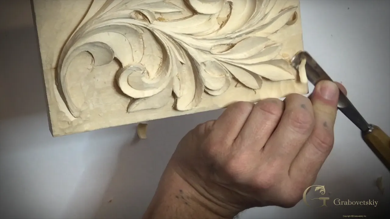 A hand drives a gouge into the curl of an acanthus leaf carved on a pale panel